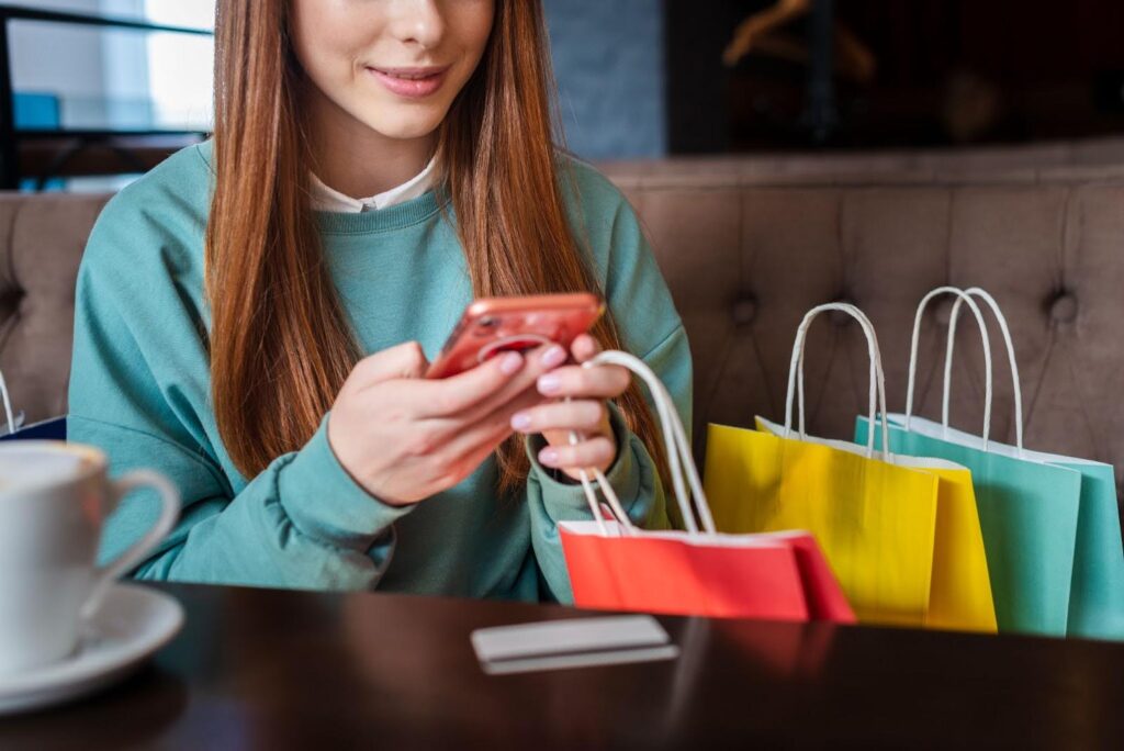 A woman sits at a table with shopping bags, holding a smartphone, browsing the best Shopify apps in the Shopify app market, with a coffee cup and credit card in front of her.
