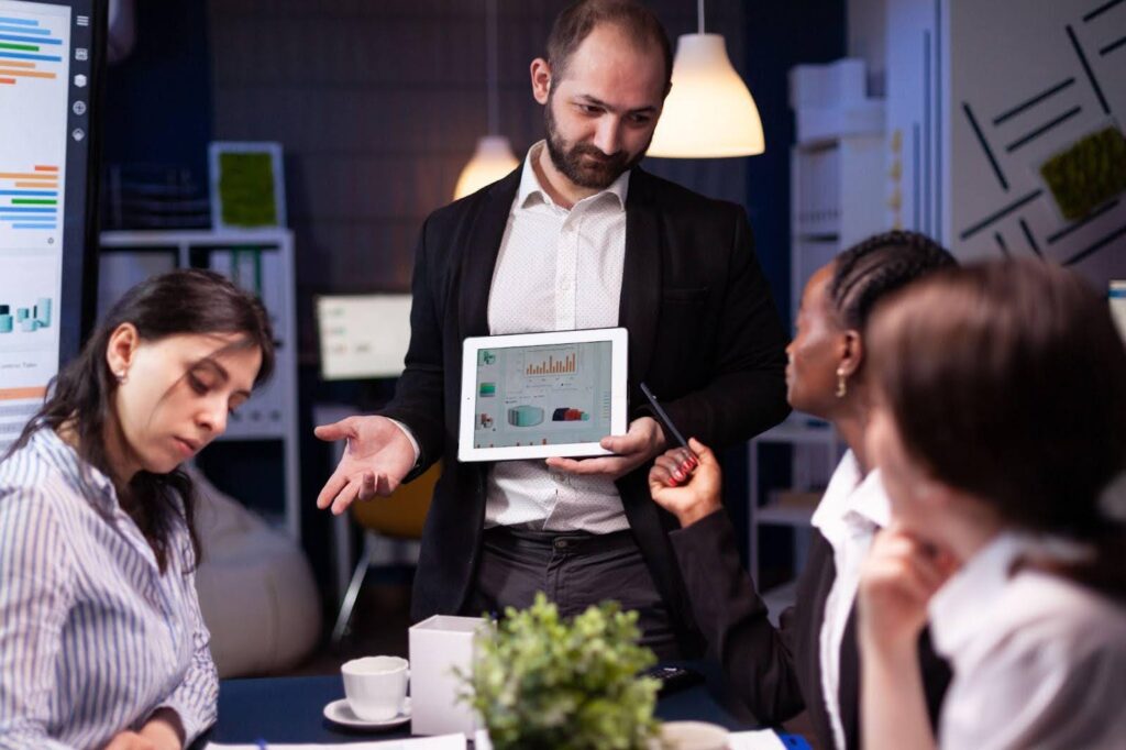 Business team collaborating around a table while a man presents data on a tablet using cloud-based productivity suites and productivity software.